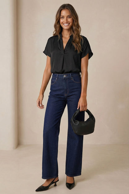 Woman wearing a drop shoulder black blouse and blue jeans holding a black handbag against a beige background