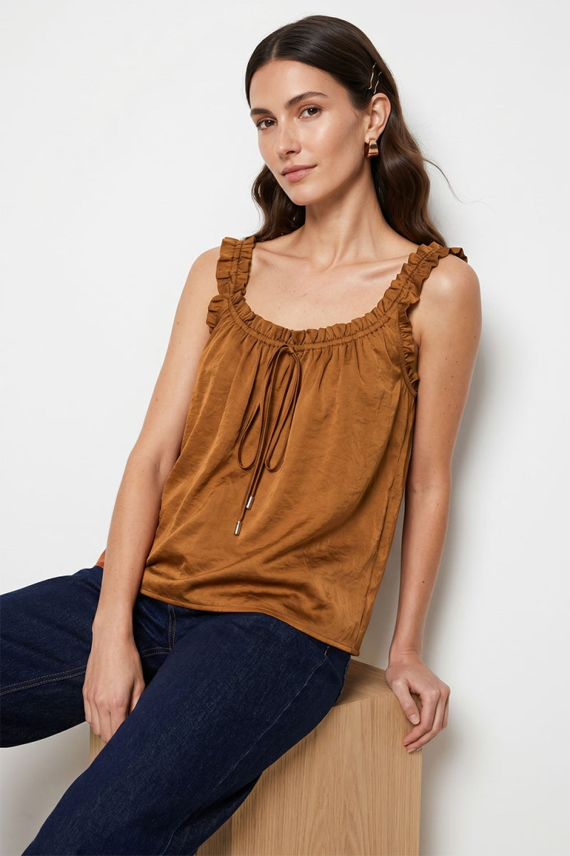 Woman wearing a rust sleeveless ruffle trim tie neck tank top with ruffled details and dark jeans, sitting on a white background.