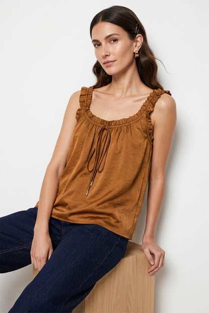 Woman wearing a rust sleeveless ruffle trim tie neck tank top with ruffled details and dark jeans, sitting on a white background.