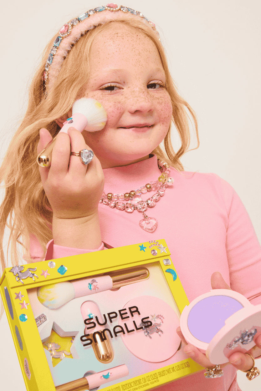 Child holding makeup products and a box labeled 'Super Smalls' against a plain background