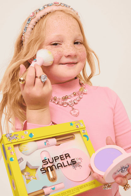 Child holding makeup products and a box labeled 'Super Smalls' against a plain background