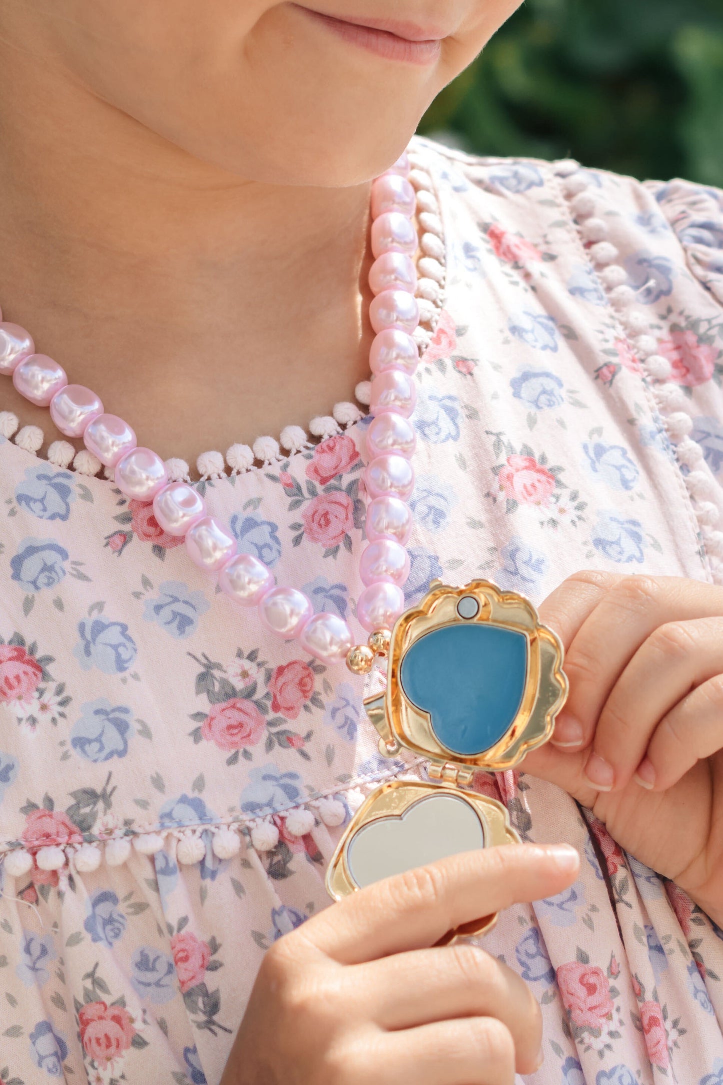 A close-up lifestyle shot of a person wearing a pink pearl beaded necklace and holding an open gold locket with a blue lip gloss center.
