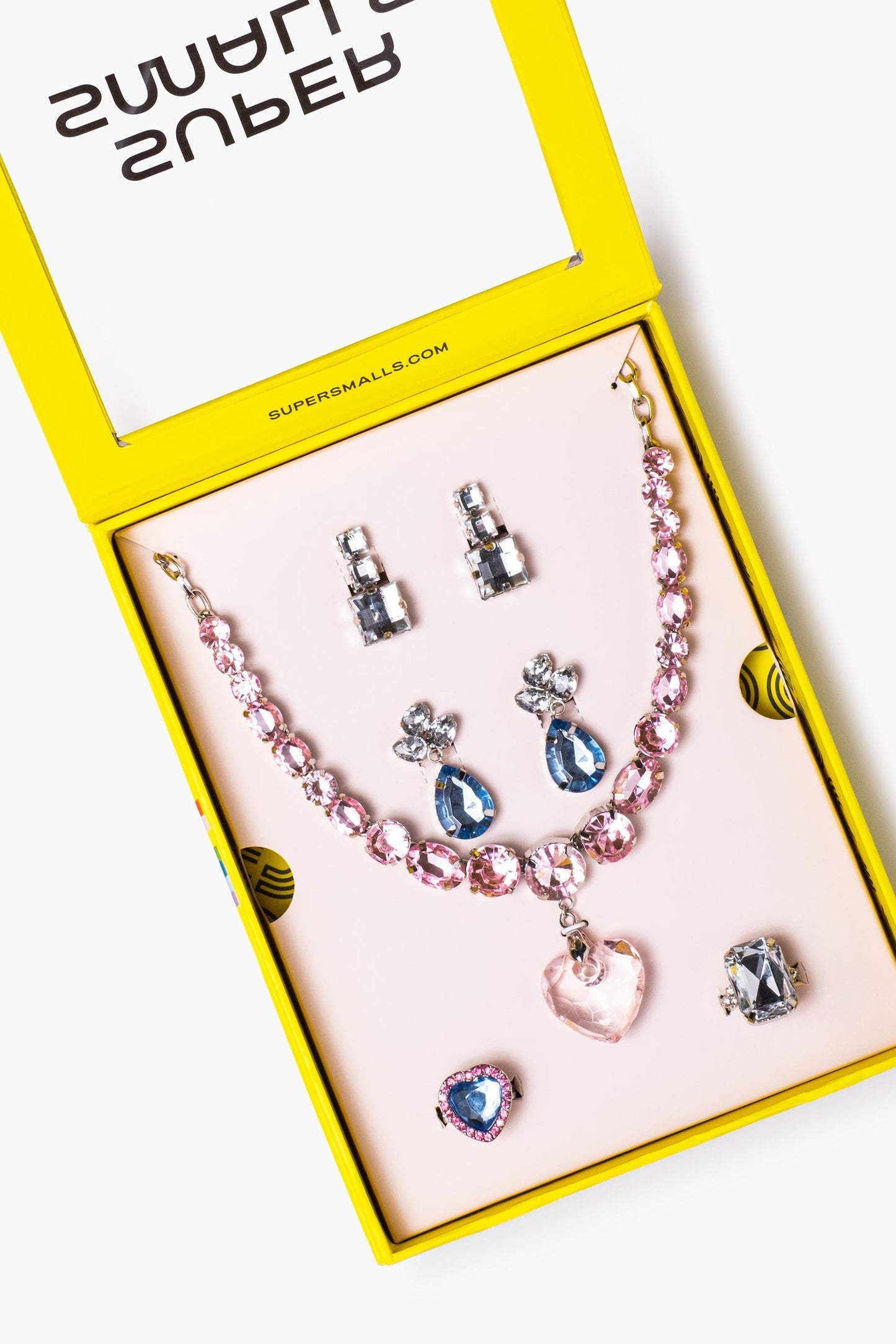 A 'Big Pres' jewelry set displayed inside an open bright yellow box on a pale pink insert. The set includes a pink beaded necklace with a heart-shaped pendant, silver and blue teardrop earrings, matching stud earrings, and a silver ring with a dark stone.