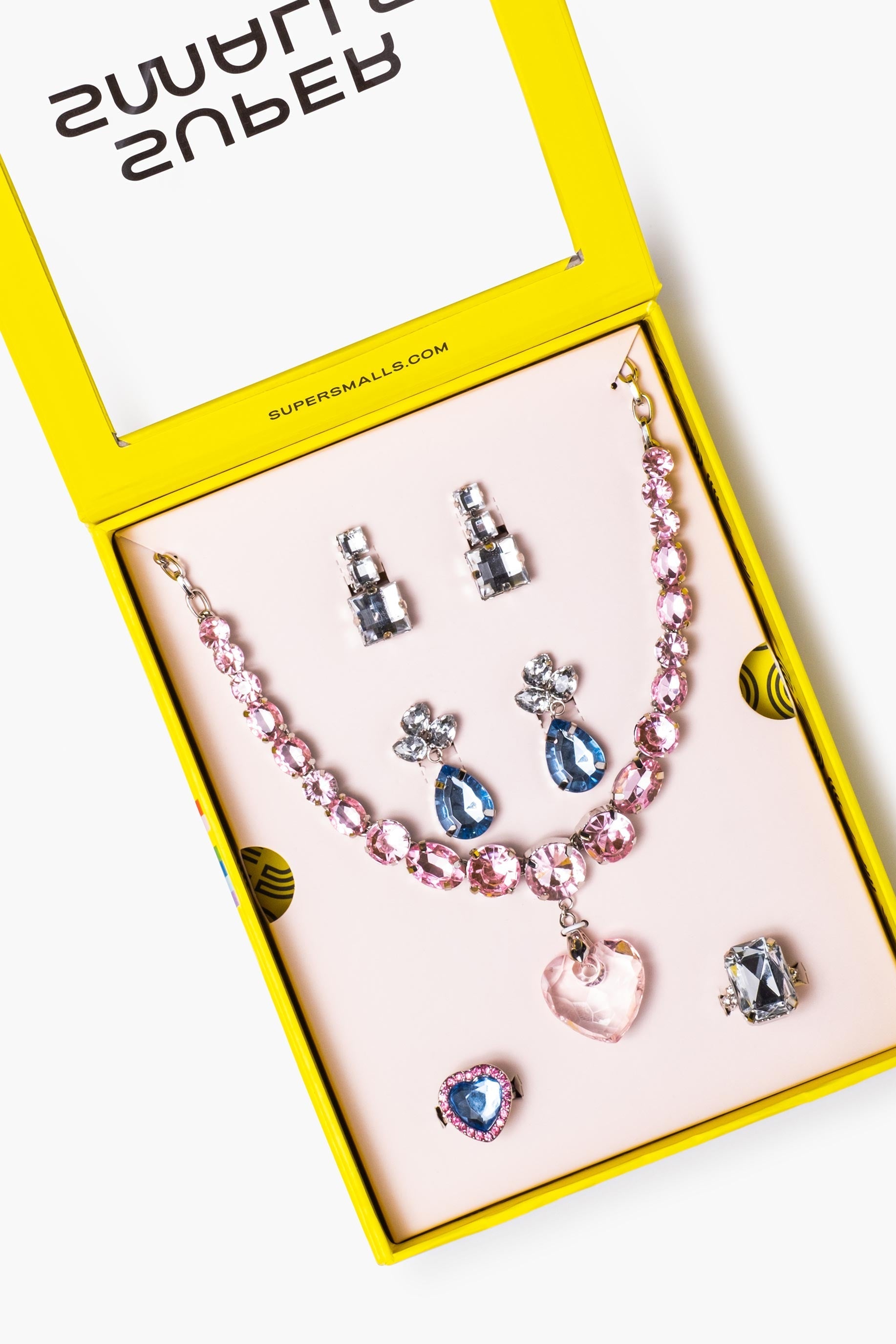 A 'Big Pres' jewelry set displayed inside an open bright yellow box on a pale pink insert. The set includes a pink beaded necklace with a heart-shaped pendant, silver and blue teardrop earrings, matching stud earrings, and a silver ring with a dark stone.