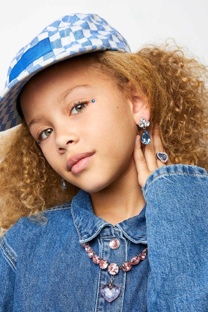 Young girl wearing a blue denim jacket, checkered hat, and 'big pres' jewelry against a white background