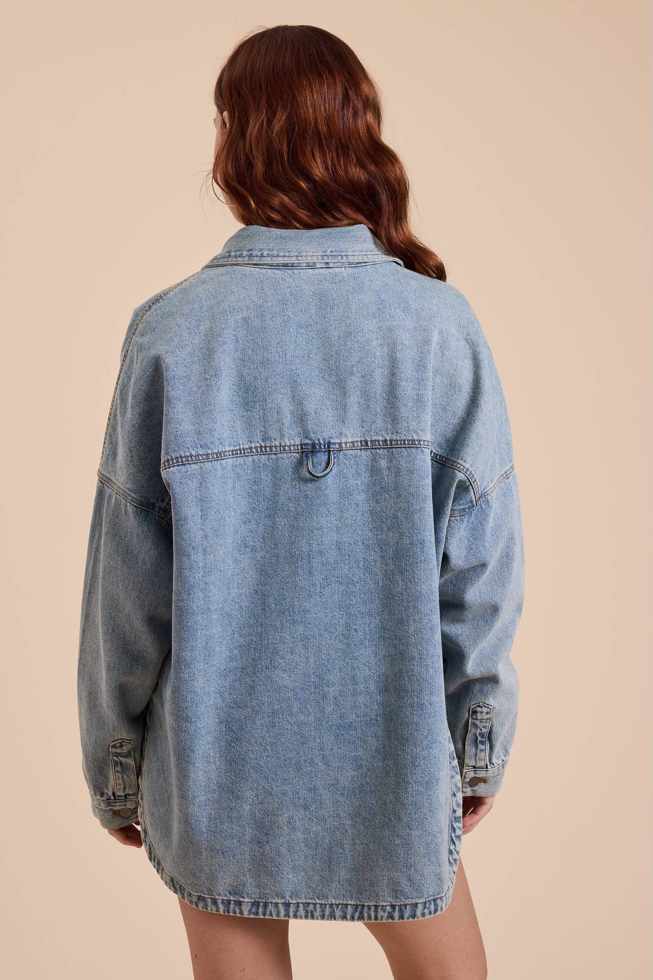 A woman viewed from the back wearing an oversized light-wash blue denim shacket set against a solid beige background.