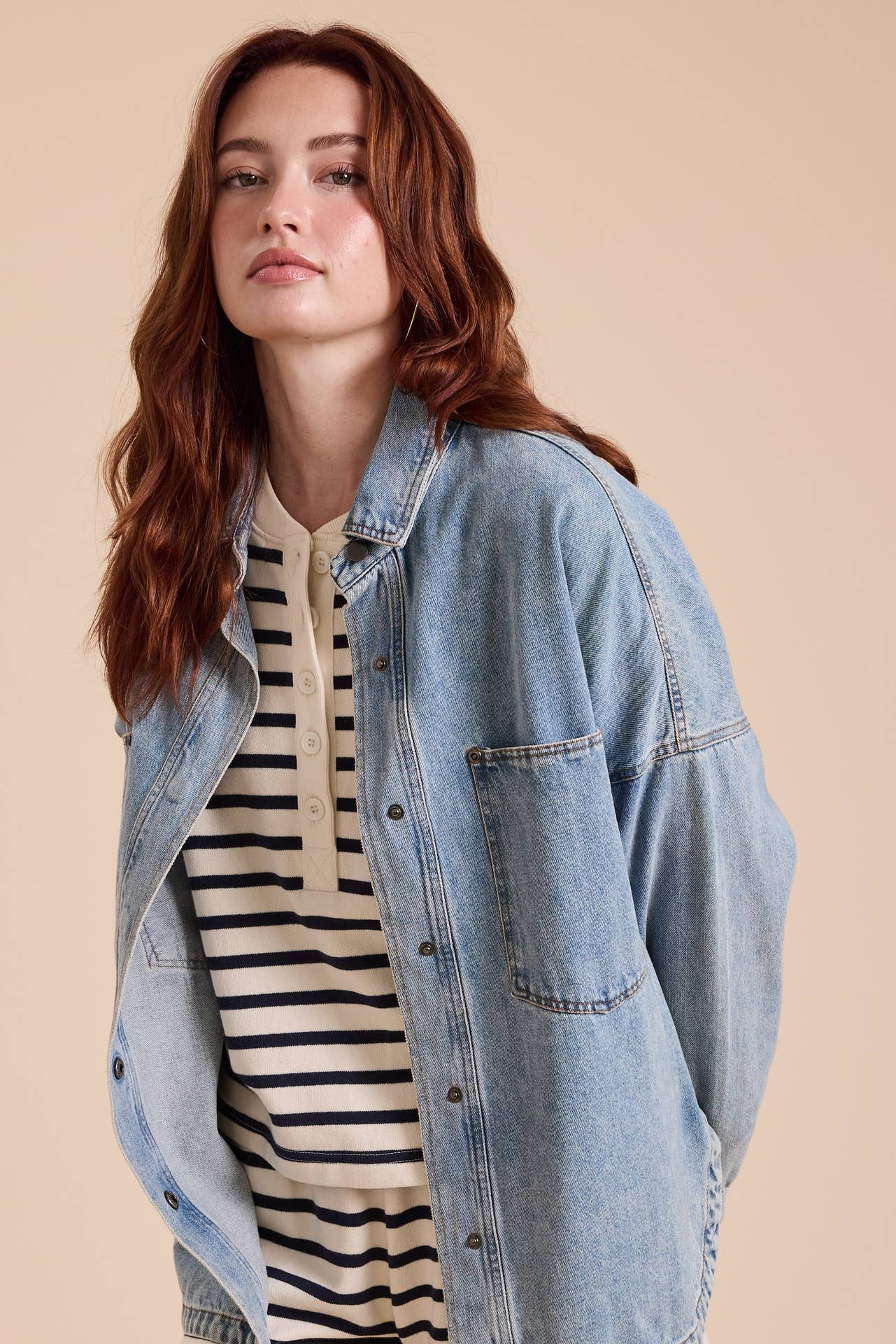 A woman with wavy auburn hair wearing a light blue denim button-up shacket over a cream and black striped henley t-shirt, posed against a solid beige studio background.