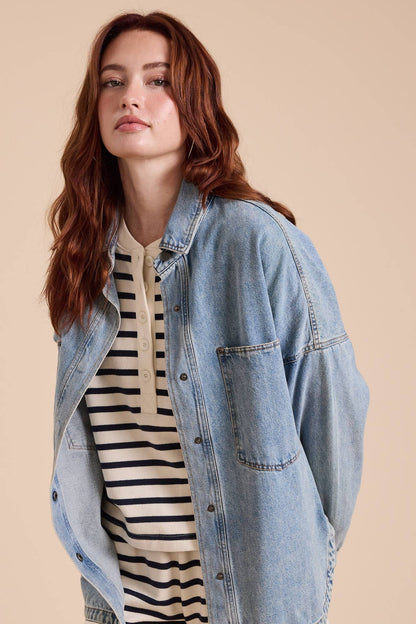 A woman with wavy auburn hair wearing a light blue denim button-up shacket over a cream and black striped henley t-shirt, posed against a solid beige studio background.