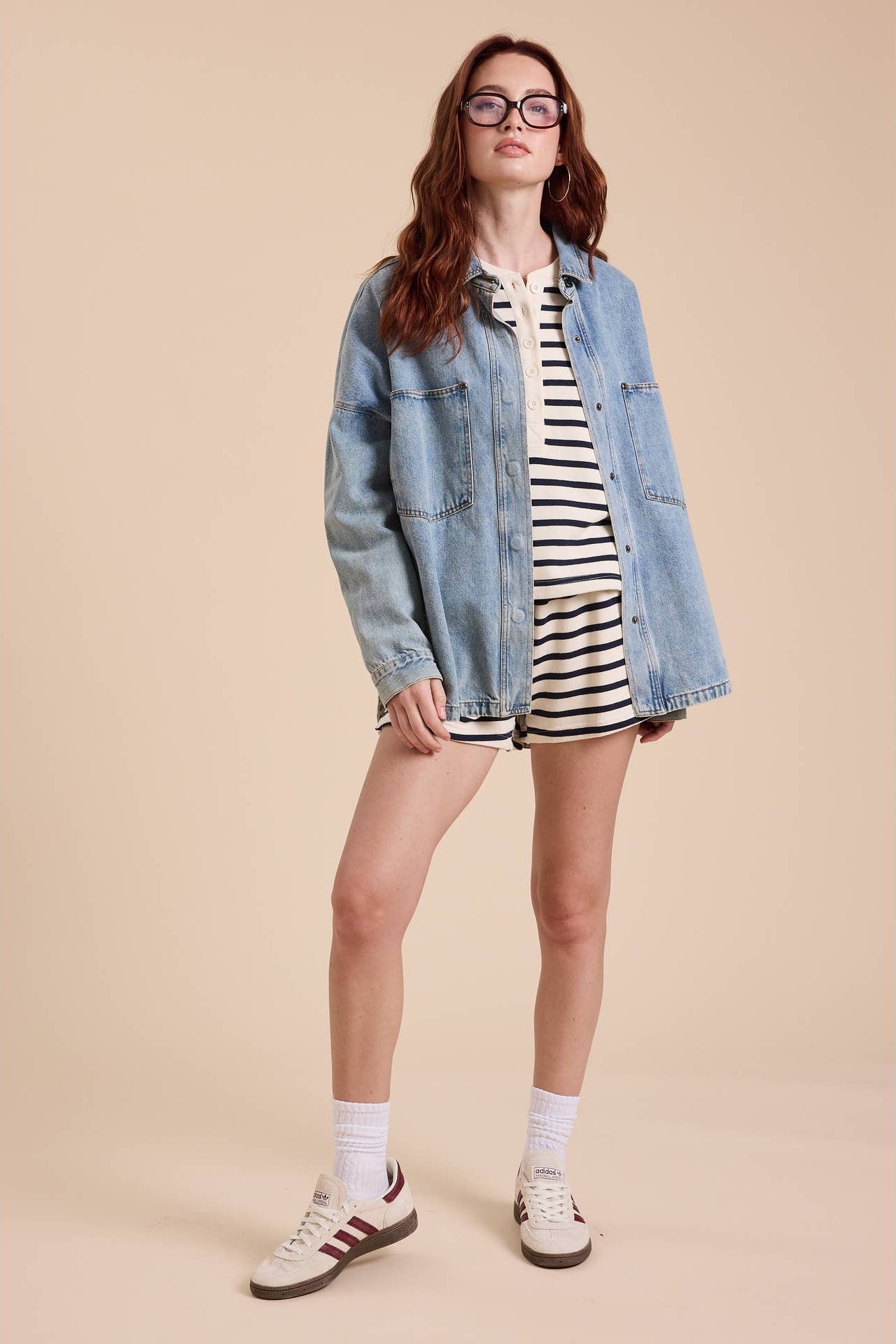 A woman modeling an oversized light blue denim shacket over a black and white striped shirt and shorts, paired with white socks and white sneakers, against a solid beige studio background.