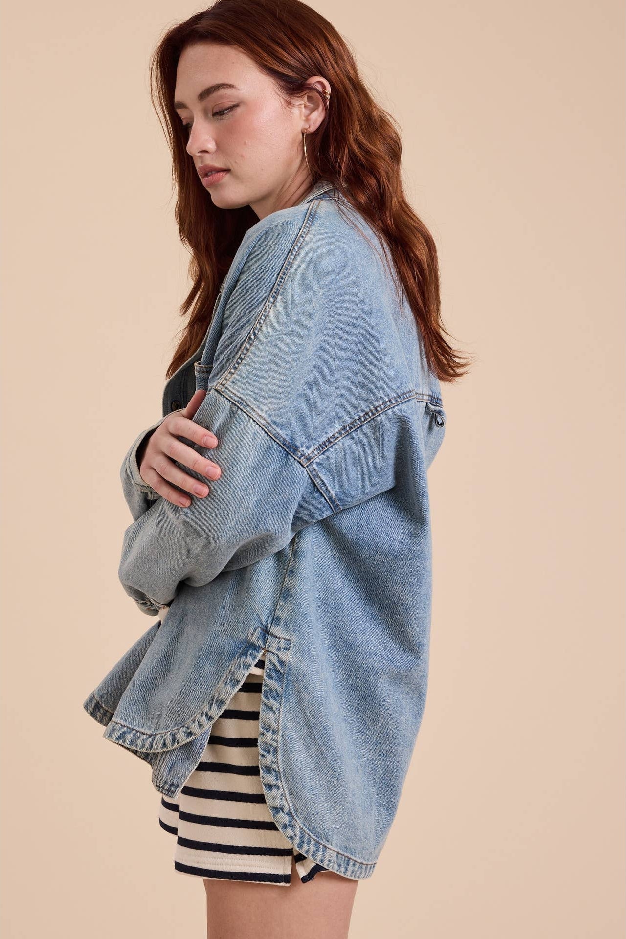 A woman wearing an oversized light-wash blue denim shacket over a black and white striped top, posed against a solid beige background.