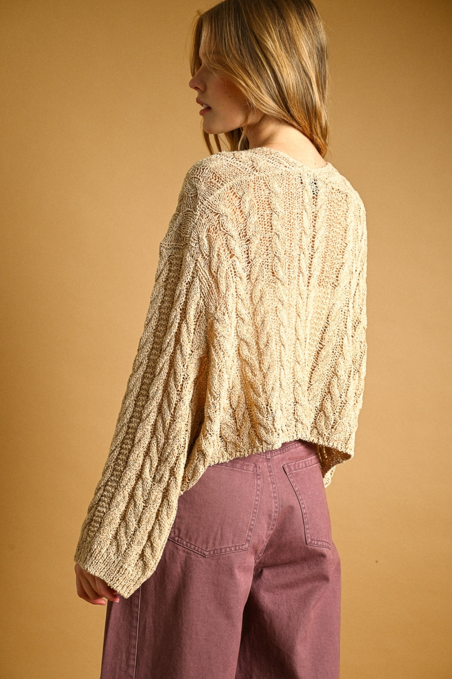 Back view of woman wearing a straw colored cable knit sweater and mauve pants against a beige background