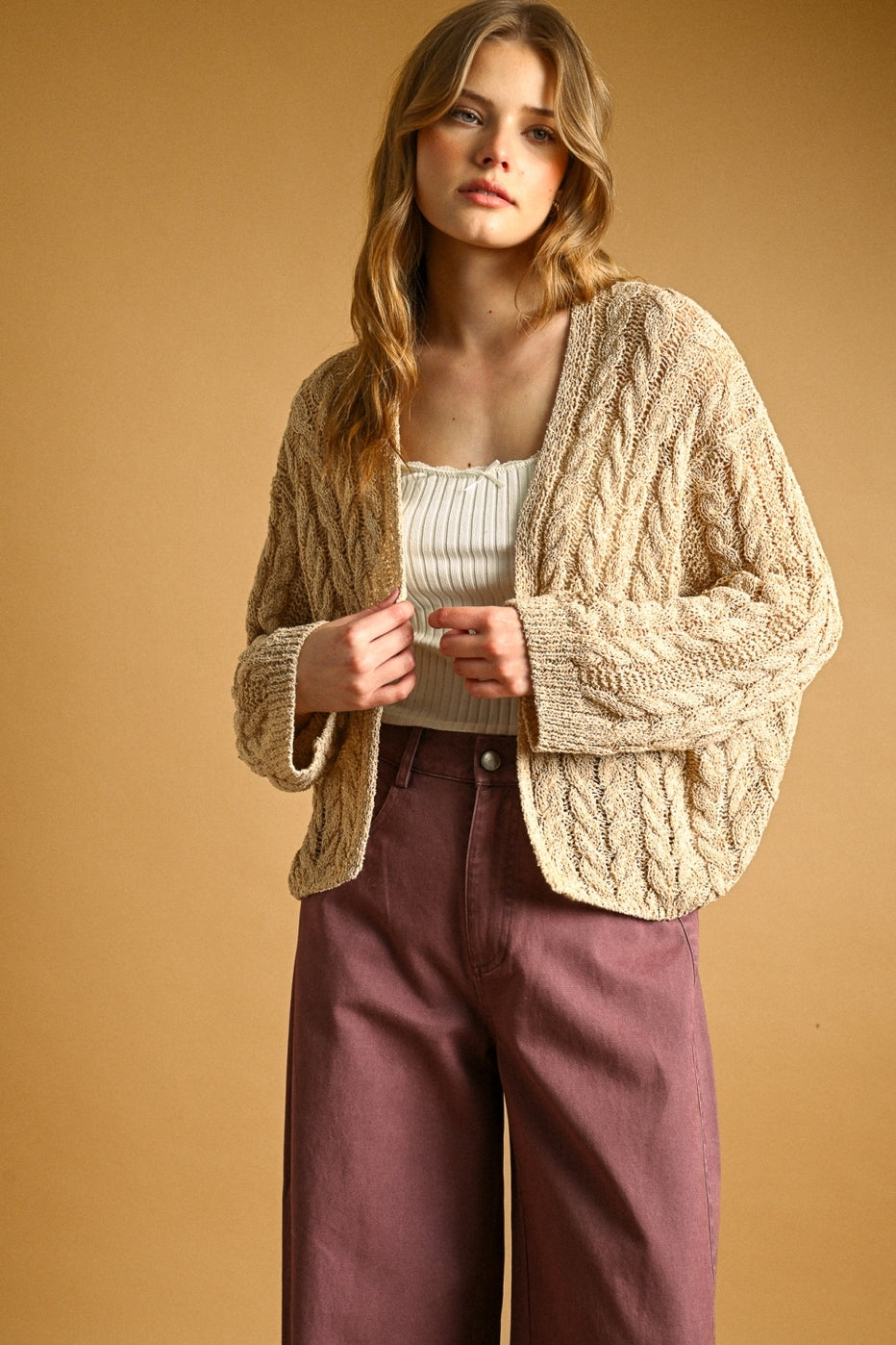 A woman modeling a chunky cable-knit cardigan in a warm straw color, worn over a cream ribbed top and mauve wide-leg pants, set against a solid warm tan studio background.