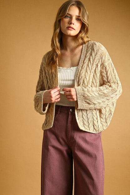 A woman modeling a chunky cable-knit cardigan in a warm straw color, worn over a cream ribbed top and mauve wide-leg pants, set against a solid warm tan studio background.