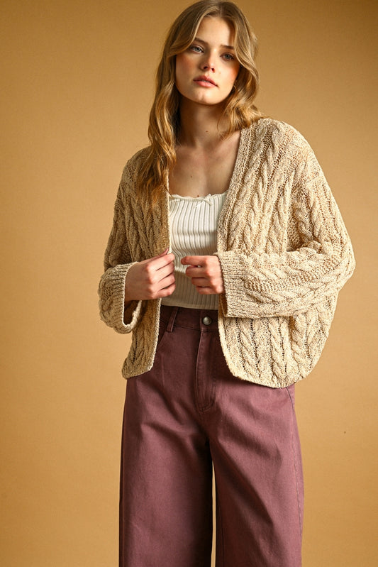 A woman modeling a chunky cable-knit cardigan in a warm straw color, worn over a cream ribbed top and mauve wide-leg pants, set against a solid warm tan studio background.