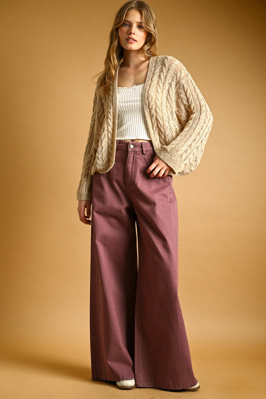 Woman wearing a straw cardigan and mauve pants against a beige background