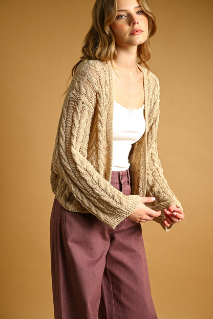 A woman wearing a chunky cable-knit straw cardigan over a white tank top and mauve wide-leg pants, posed against a solid warm tan studio background.
