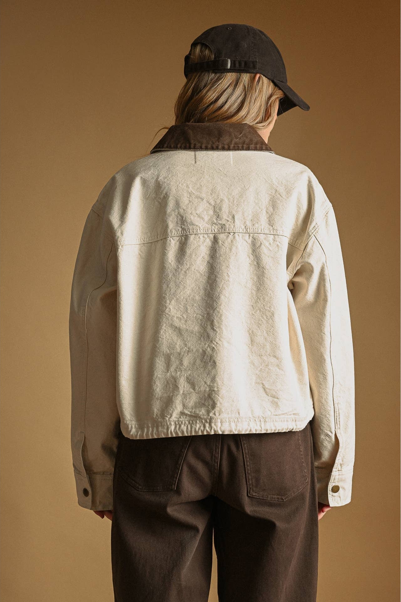 A person shown from the back wearing an ecru cropped utility jacket with a dark brown corduroy collar, dark brown pants, and a black baseball cap, set against a solid warm brown background.