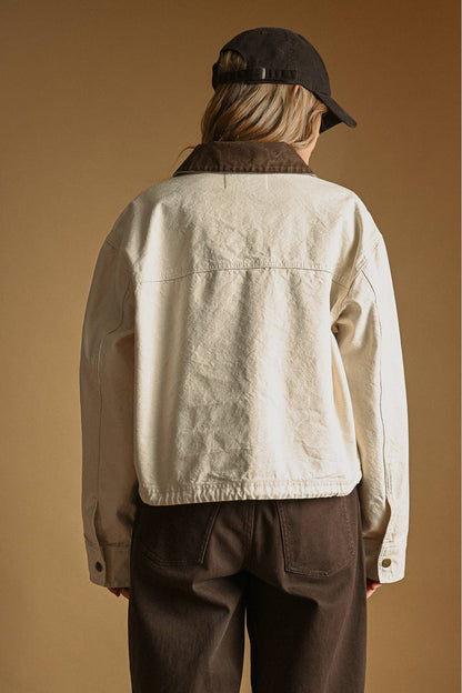 A person shown from the back wearing an ecru cropped utility jacket with a dark brown corduroy collar, dark brown pants, and a black baseball cap, set against a solid warm brown background.