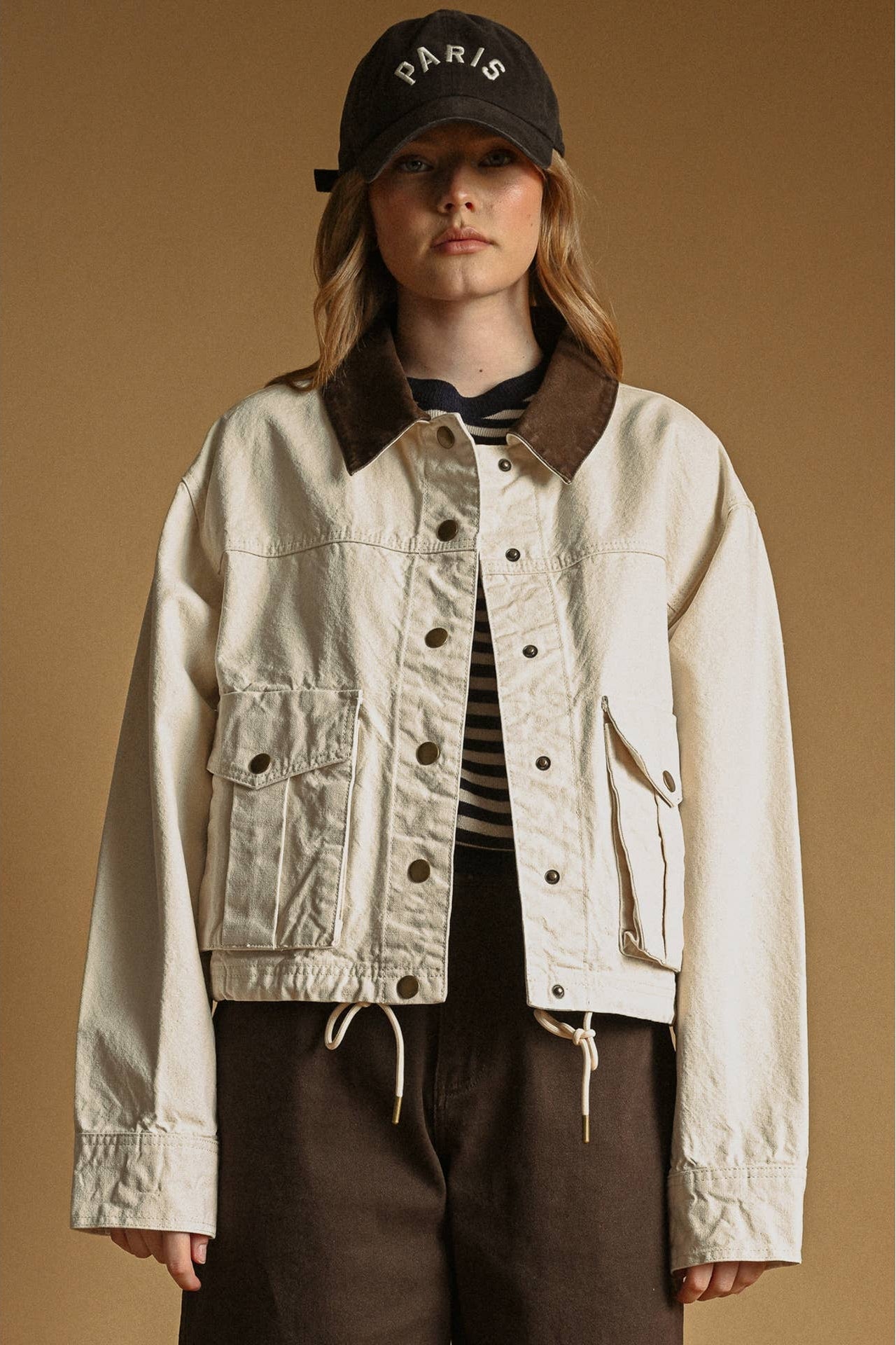 A model wearing an ecru-colored utility jacket with a brown corduroy collar, styled with a black cap, striped shirt, and brown pants against a solid tan background.