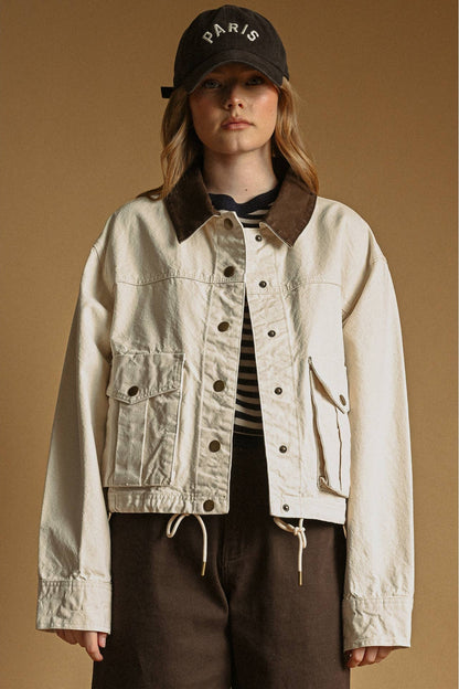 A model wearing an ecru-colored utility jacket with a brown corduroy collar, styled with a black cap, striped shirt, and brown pants against a solid tan background.