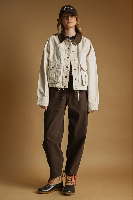 A model stands against a warm tan studio background wearing an ecru colored cropped utility jacket with dark brown corduroy collar details, dark brown pants, brown and black duck boots, and a black baseball cap.
