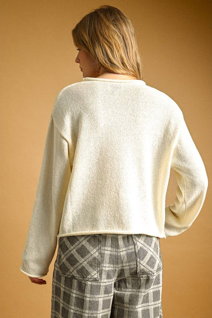 A woman shown from the back wearing a cream-colored, cropped, long-sleeve knit sweater and gray plaid pants, set against a solid warm tan background.