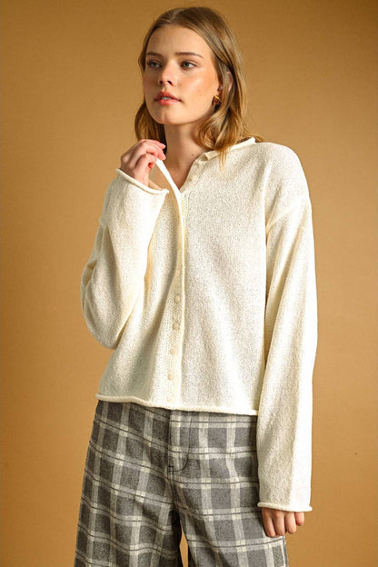 A woman modeling a cream-colored, textured long-sleeve button-up cardigan paired with gray and white plaid trousers, set against a solid warm brown background.