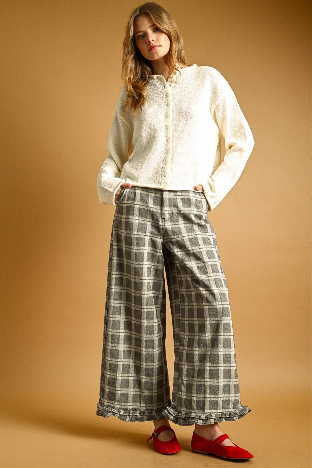 Woman wearing a cream button up cardigan with plaid brown and cream pants and red shoes against a light brown background