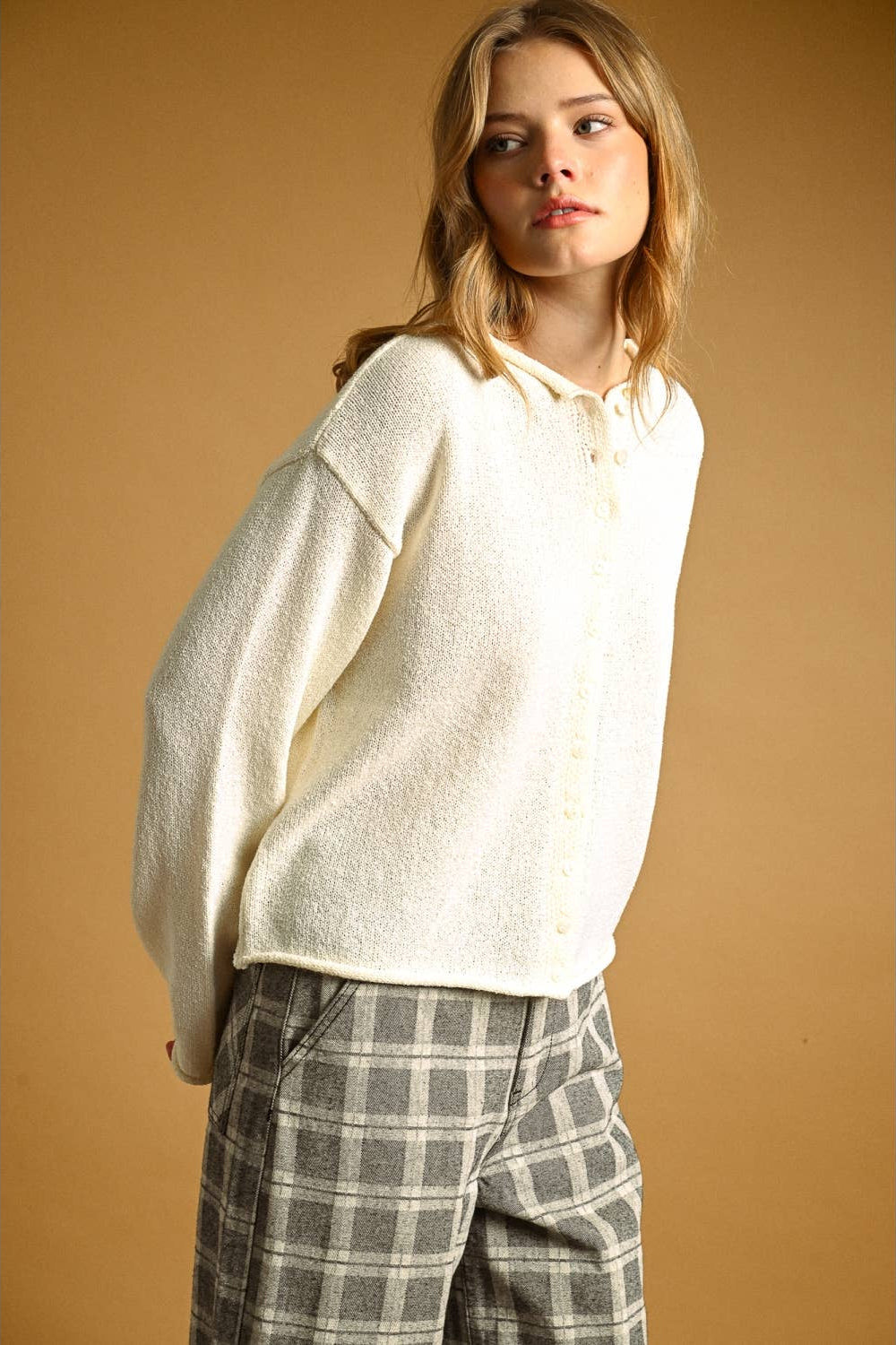 A woman wearing a cream-colored, oversized  knit button-up cardigan and gray-and-white plaid trousers, standing against a solid warm brown studio background.