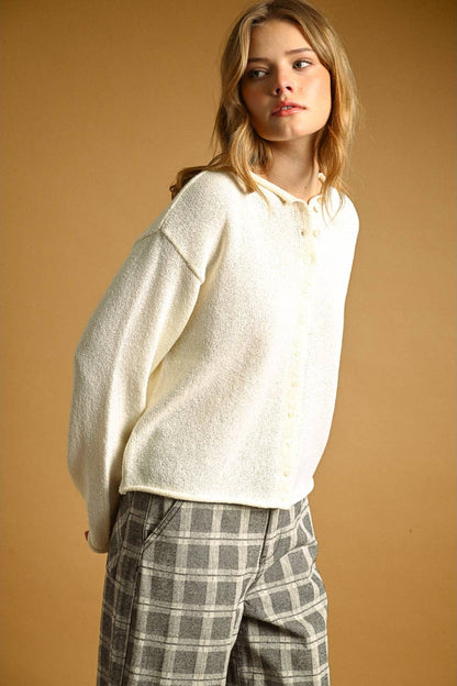 A woman wearing a cream-colored, oversized  knit button-up cardigan and gray-and-white plaid trousers, standing against a solid warm brown studio background.