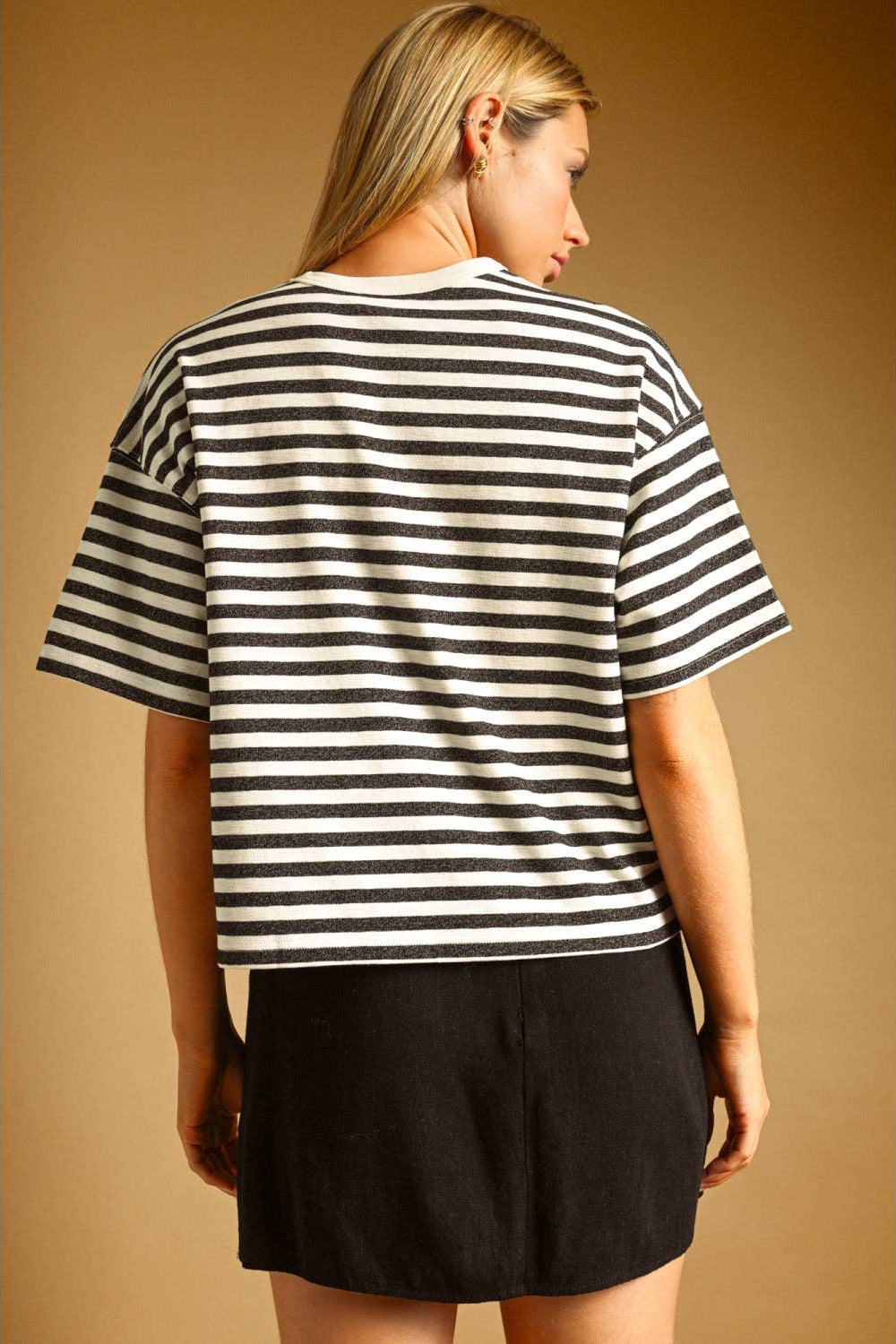 A woman shown from the back wearing a charcoal and white horizontally striped short-sleeve t-shirt and black shorts, set against a solid warm brown studio background.