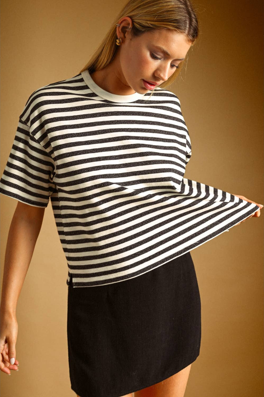 A woman wearing a charcoal and white horizontally striped short-sleeve knit top and a black skirt, posing against a warm brown studio background.