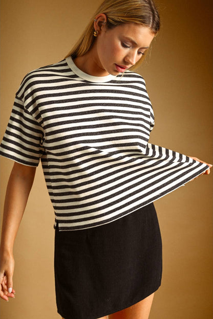 A woman wearing a charcoal and white horizontally striped short-sleeve knit top and a black skirt, posing against a warm brown studio background.