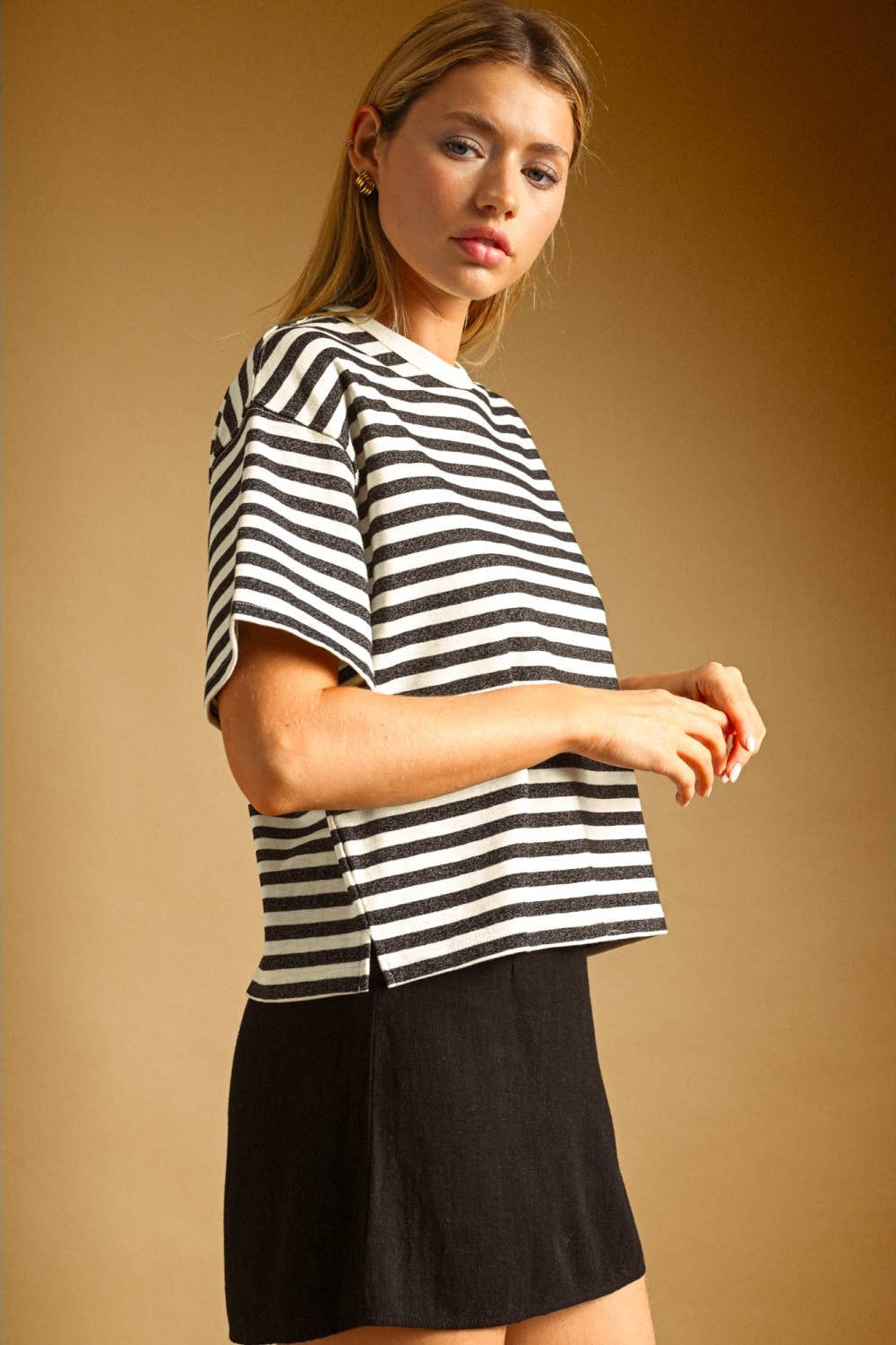 A woman wearing a charcoal and white horizontally striped short-sleeve top and a black skirt, posed against a warm tan studio background.