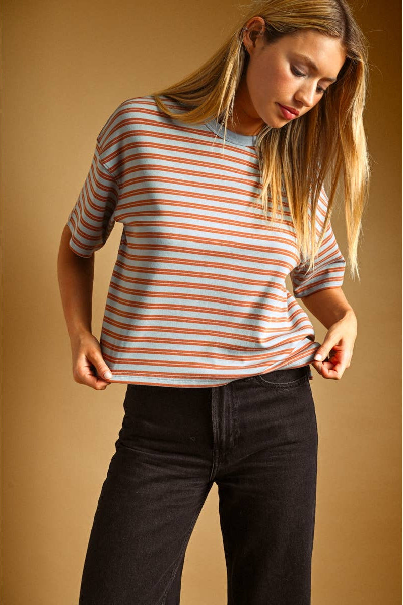 A woman wearing a short-sleeve crewneck t-shirt with thin blue and rust horizontal stripes, paired with black jeans, set against a solid warm brown studio background.