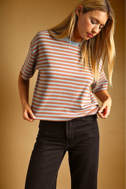 A woman wearing a short-sleeve crewneck t-shirt with thin blue and rust horizontal stripes, paired with black jeans, set against a solid warm brown studio background.