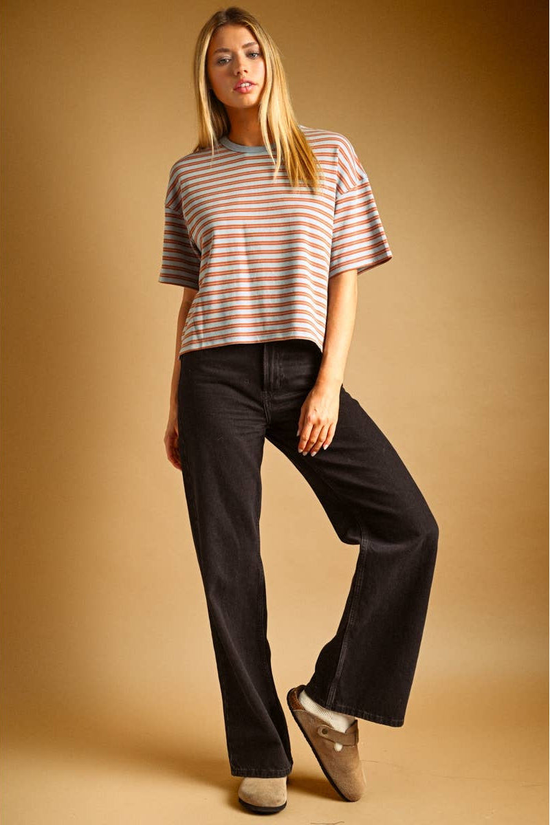 A woman modeling a short-sleeve striped knit top in blue/rust color, paired with black wide-leg jeans and tan slip-on shoes, against a warm tan studio background.