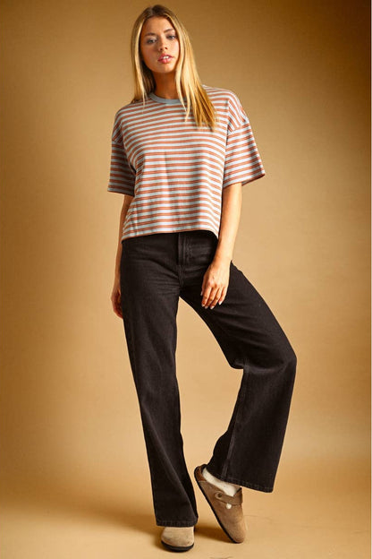 A woman modeling a short-sleeve striped knit top in blue/rust color, paired with black wide-leg jeans and tan slip-on shoes, against a warm tan studio background.