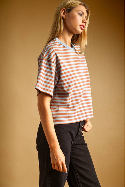 A woman wearing a short-sleeve crewneck t-shirt with thin blue and rust horizontal stripes, paired with black jeans, set against a solid warm brown studio background.