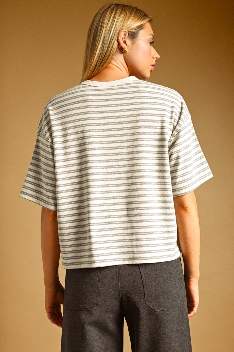 A woman shown from the back wearing a short-sleeve oat and grey striped knit top with dark gray pants, set against a solid warm tan studio background.