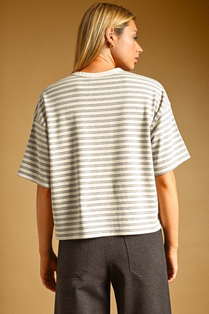 A woman shown from the back wearing a short-sleeve oat and grey striped knit top with dark gray pants, set against a solid warm tan studio background.
