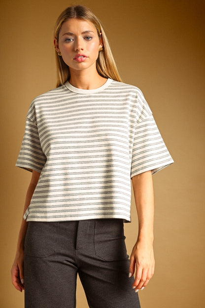 A woman modeling a short-sleeve crewneck t-shirt with thin horizontal grey stripes on an oat colored base, paired with dark gray pants, against a solid warm brown studio background.