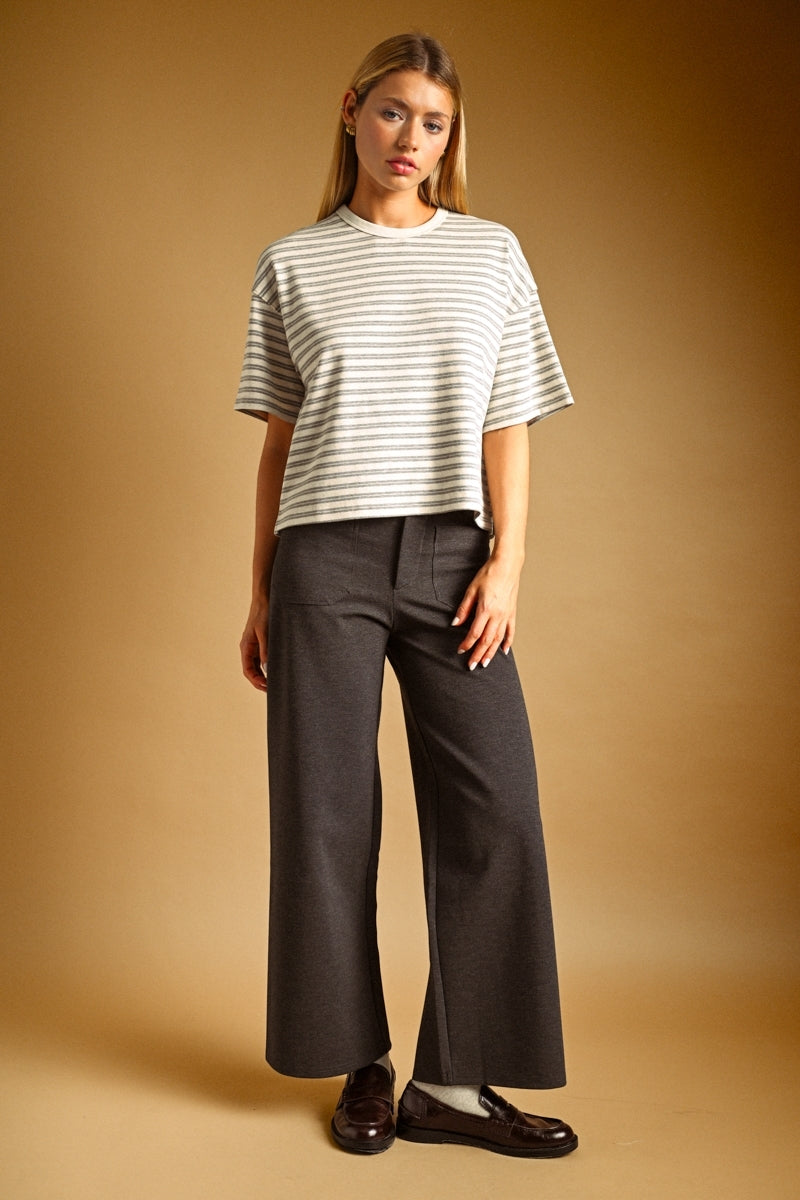A woman modeling a short-sleeve grey and oat striped knit top and dark wide-leg trousers against a warm brown studio backdrop.