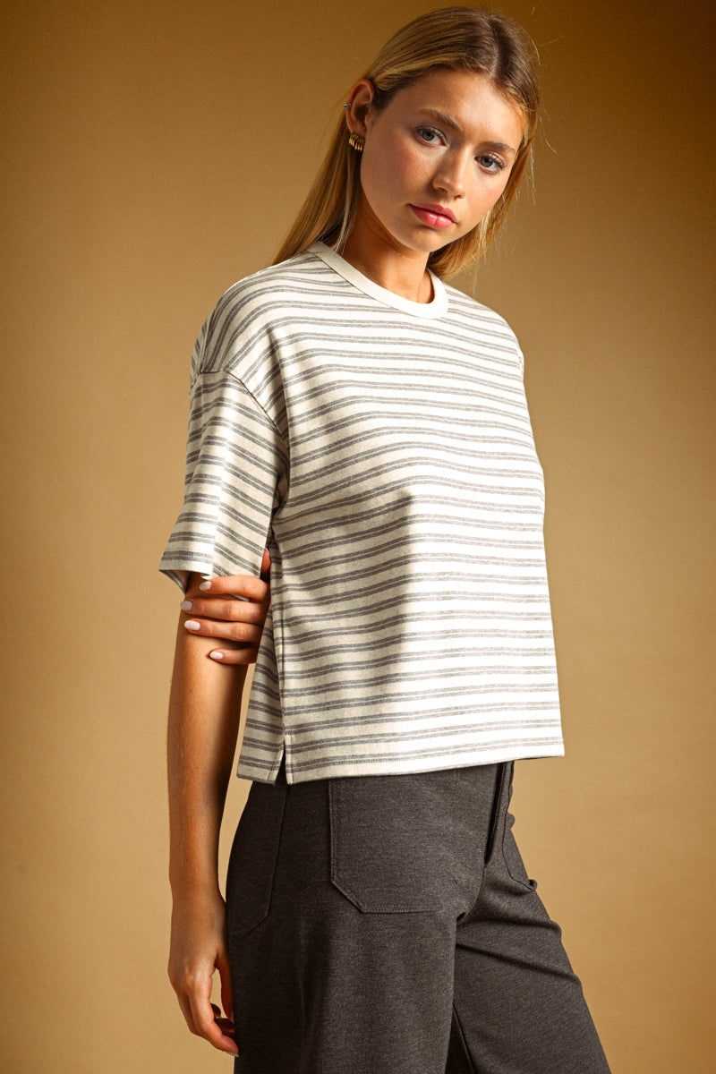 A woman wearing a short-sleeve crewneck t-shirt with thin horizontal grey stripes on an oat colored base, paired with dark gray trousers, set against a solid warm brown studio background.