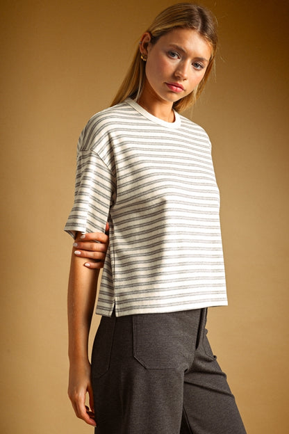 A woman wearing a short-sleeve crewneck t-shirt with thin horizontal grey stripes on an oat colored base, paired with dark gray trousers, set against a solid warm brown studio background.