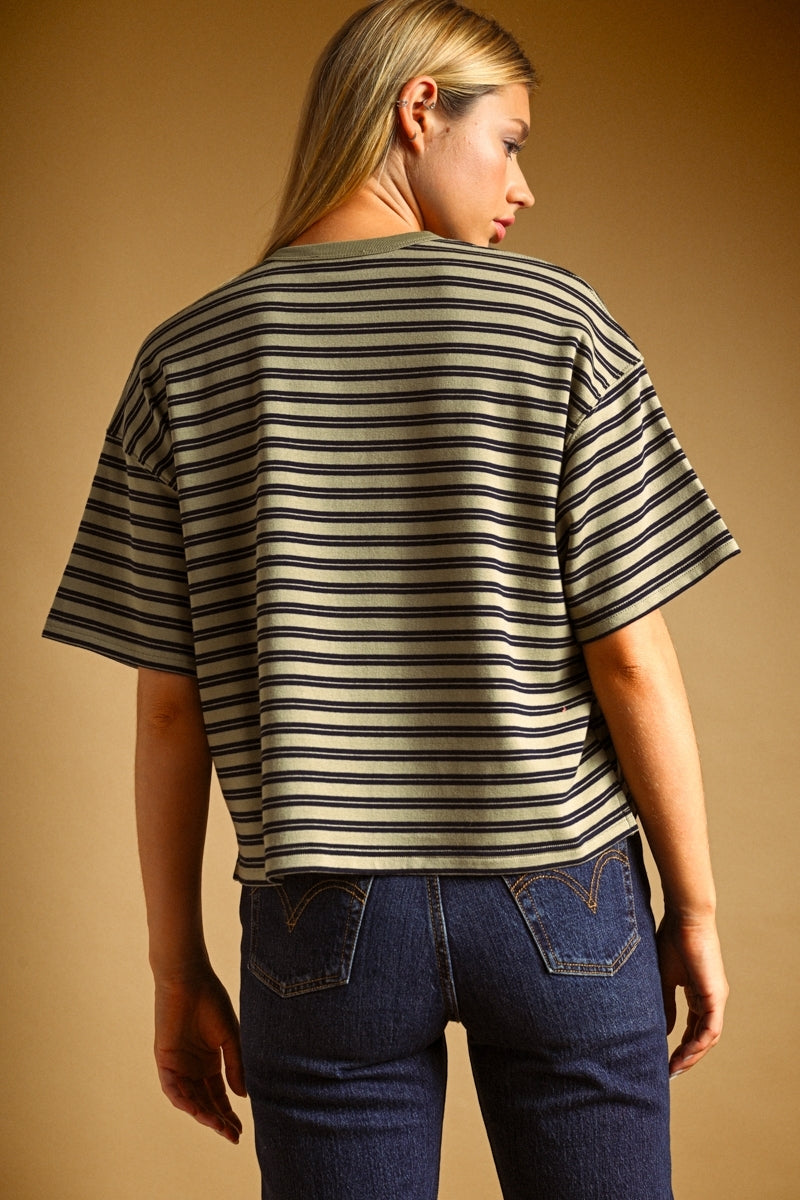 A woman shown from the back wearing a short-sleeve striped t-shirt with dark navy and olive horizontal stripes, paired with dark blue jeans, against a solid warm brown studio background.