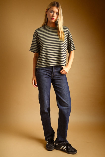 A woman modeling a short-sleeve striped t-shirt in olive green and navy, paired with dark blue jeans and black sneakers, against a warm brown studio backdrop.
