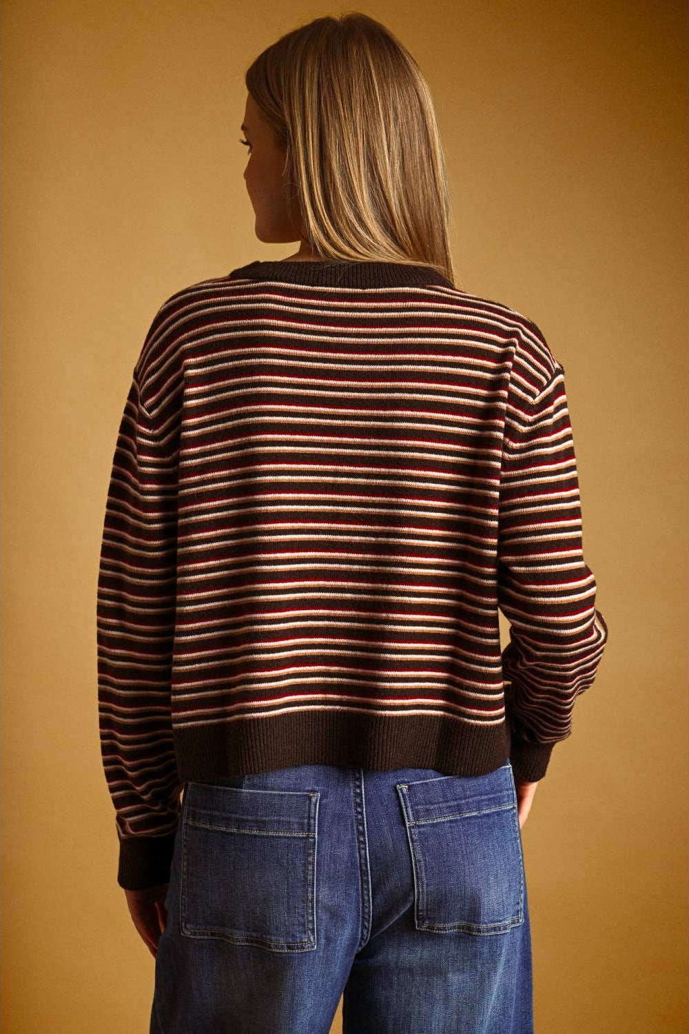 A woman viewed from the back wearing a brown knit sweater with thin horizontal red and cream stripes, paired with blue jeans, against a solid warm brown background.