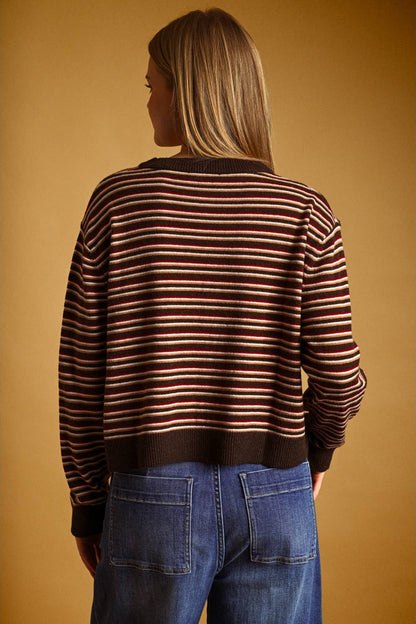 A woman viewed from the back wearing a brown knit sweater with thin horizontal red and cream stripes, paired with blue jeans, against a solid warm brown background.