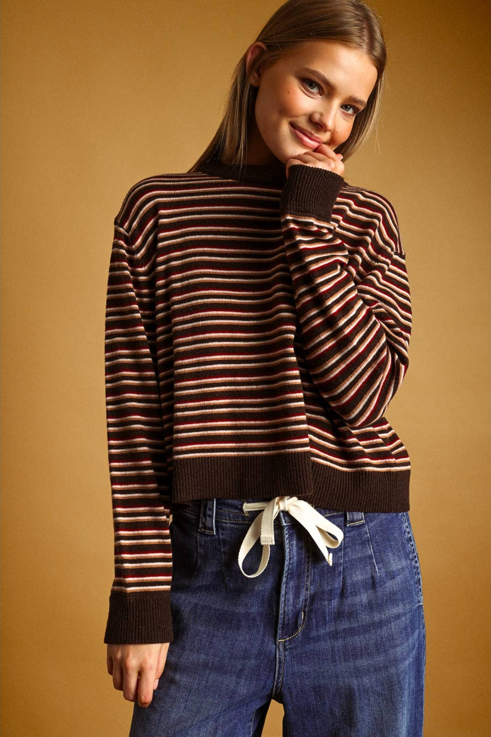 A woman wearing a brown and multi striped knit sweater with dark brown ribbed trim, paired with blue jeans featuring a white drawstring, set against a solid warm brown background.